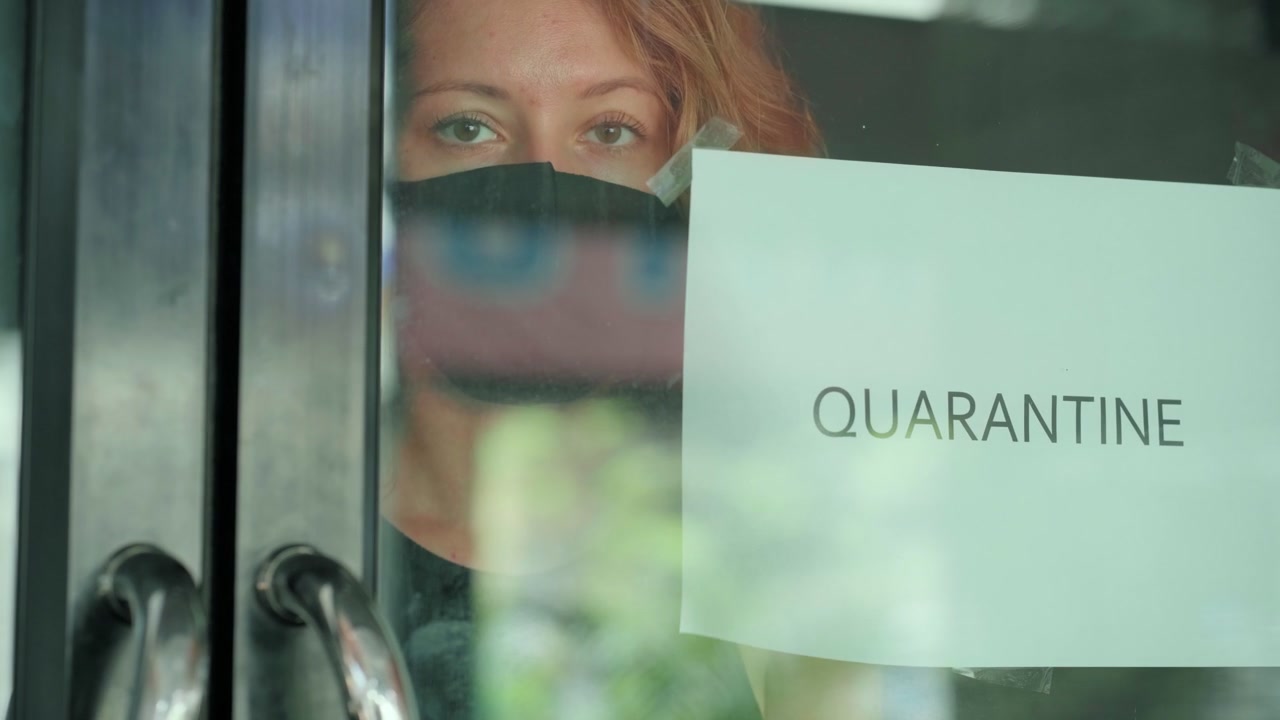 Donna che indossa una mascherina protettiva e guarda attraverso una porta a vetri con un cartello di quarantena