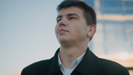 business man in black clothing gazes into distant with a warm smile, light illuminating his left shoulder, background features modern urban building