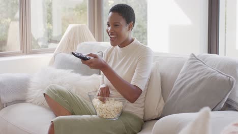 happy african american woman sitting on sofa, watching tv and eating popcorn, slow motion