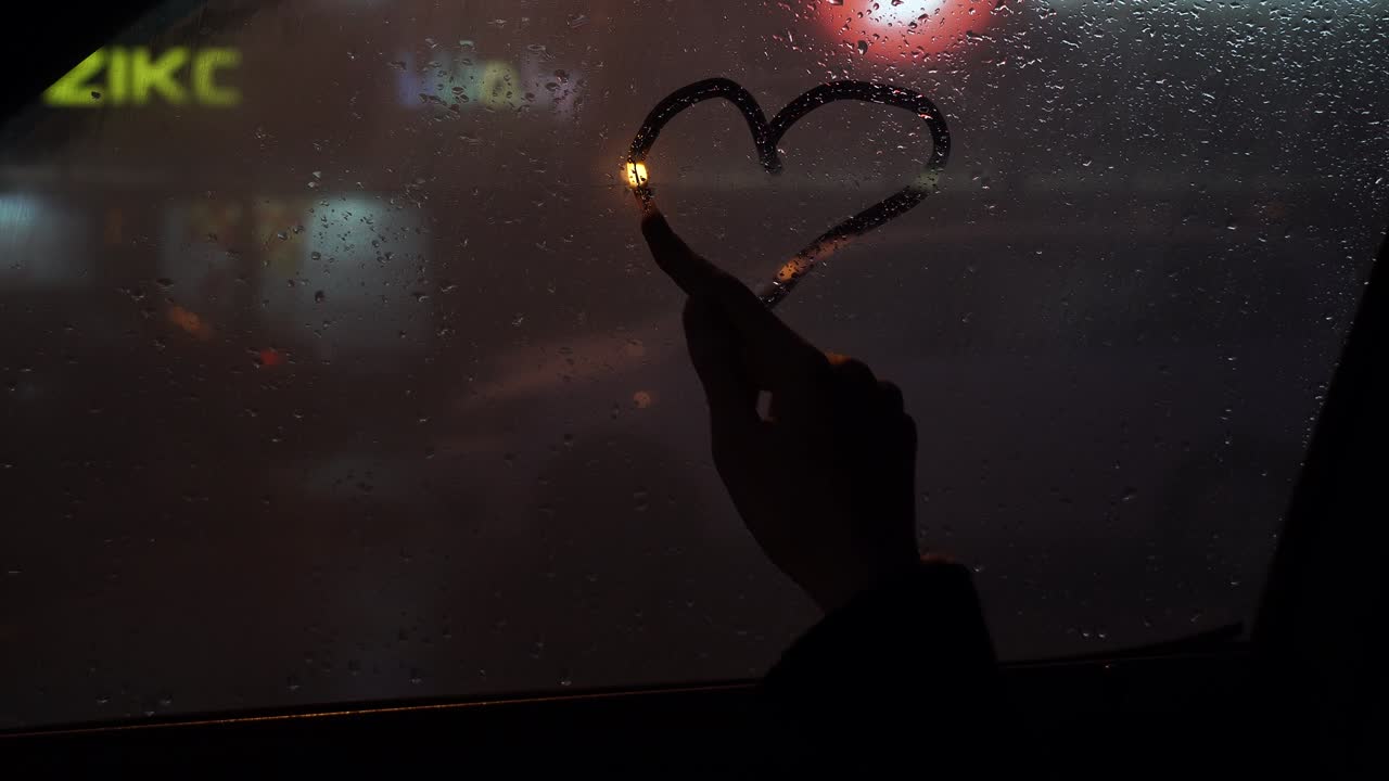 창문의 유리창이 물에 잠겼다: 지표손가락을 가진 어린 소녀는 사랑과 마음의 상징을 그린다 - 성 발렌타인 데이