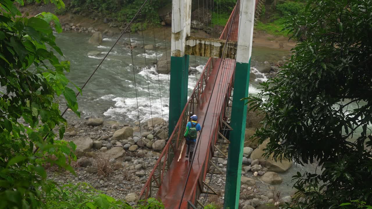 Shot from a forest-side angle, a hiker cross a narrow suspension bridge over a rapid Philippine river, framed by lush greenery and rugged terrain