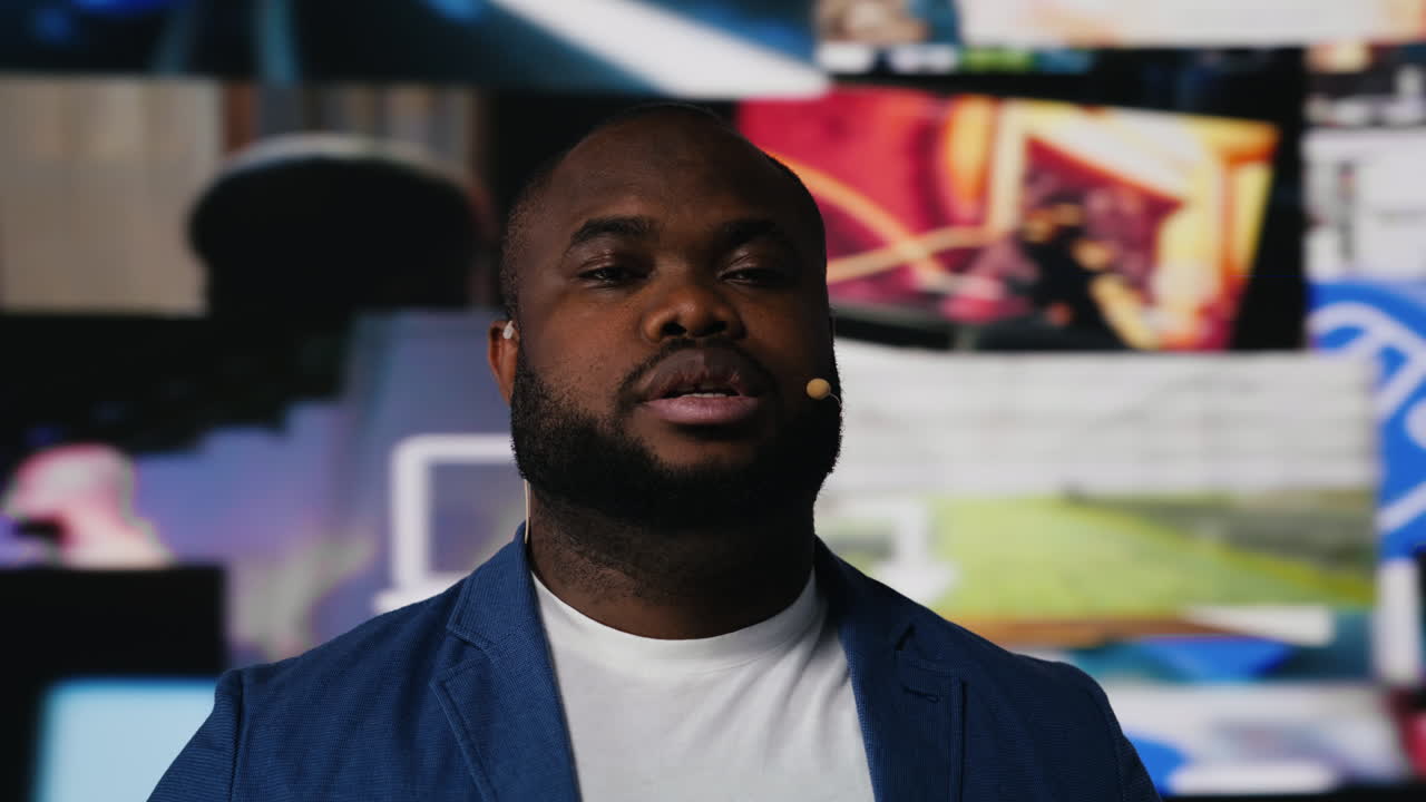 Black web coach hosting a discussion on stage surrounded by media visuals