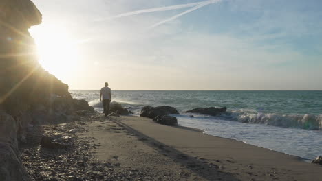 man walks along beach during sunny morning, long shadow casts behind, sea waves wash onshore