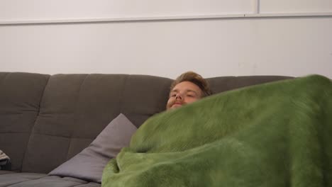 A-blonde-guy-with-a-smart-mustache,-who-is-wrapping-himself-in-his-green-blanket-for-watching-the-TV