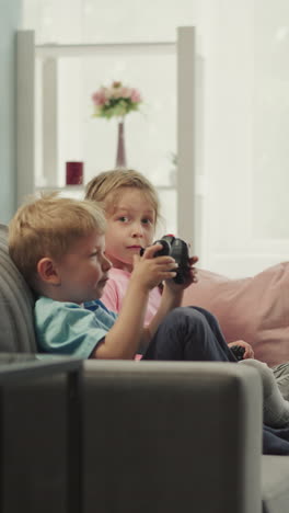 cute toddler boy learns to use gaming console with elder sister sitting on sofa at home. siblings enjoy playing game together after classes