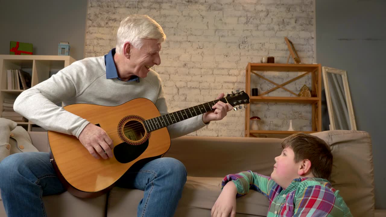 el abuelo toca para su nieto la guitarra, la música. un joven gordo está sentado en el refugio con su abuelo. enseña a su nieto a tocar la guitarra. confort en el hogar, idilio familiar, concepto de acogida, diferencia de generaciones. 60 fps