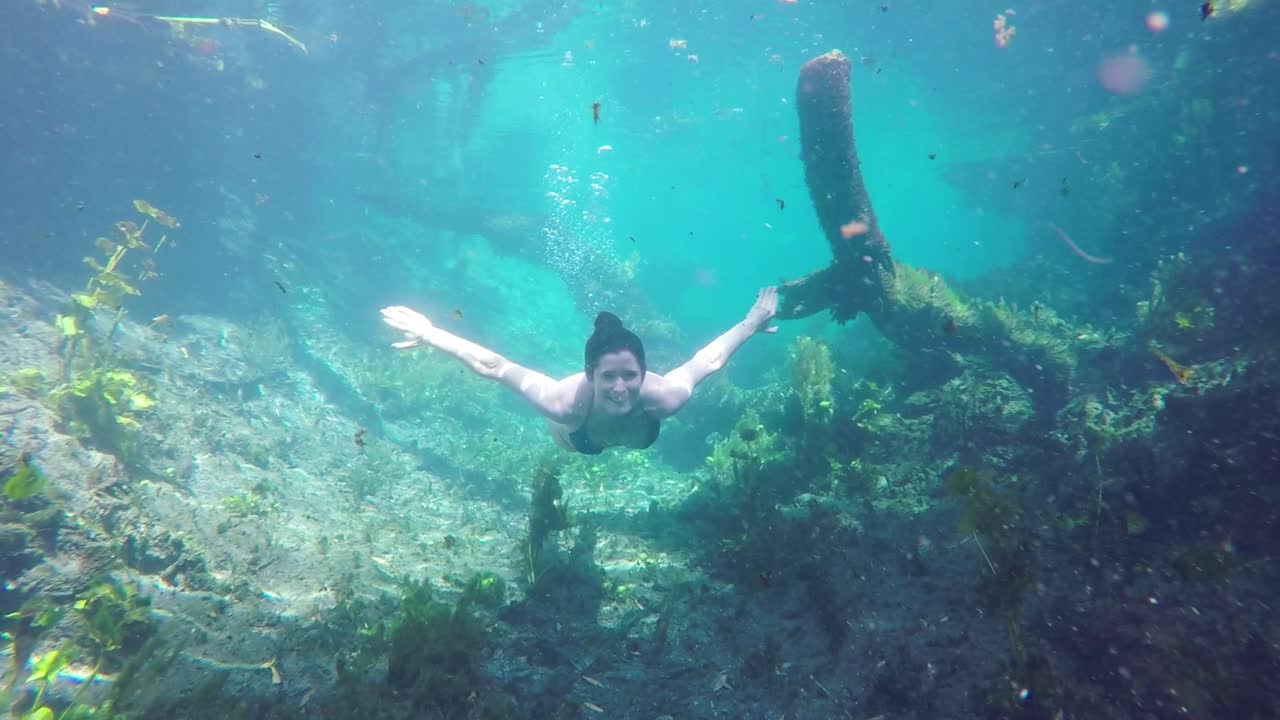 맑고 푸른 물 속에서 수영하는 비키니 입은 여성의 슬로우 모션, 그녀가 카메라에 접근할 때 손을 흔들고 미소를 지으며 수면 위로 떠오르기 시작합니다.