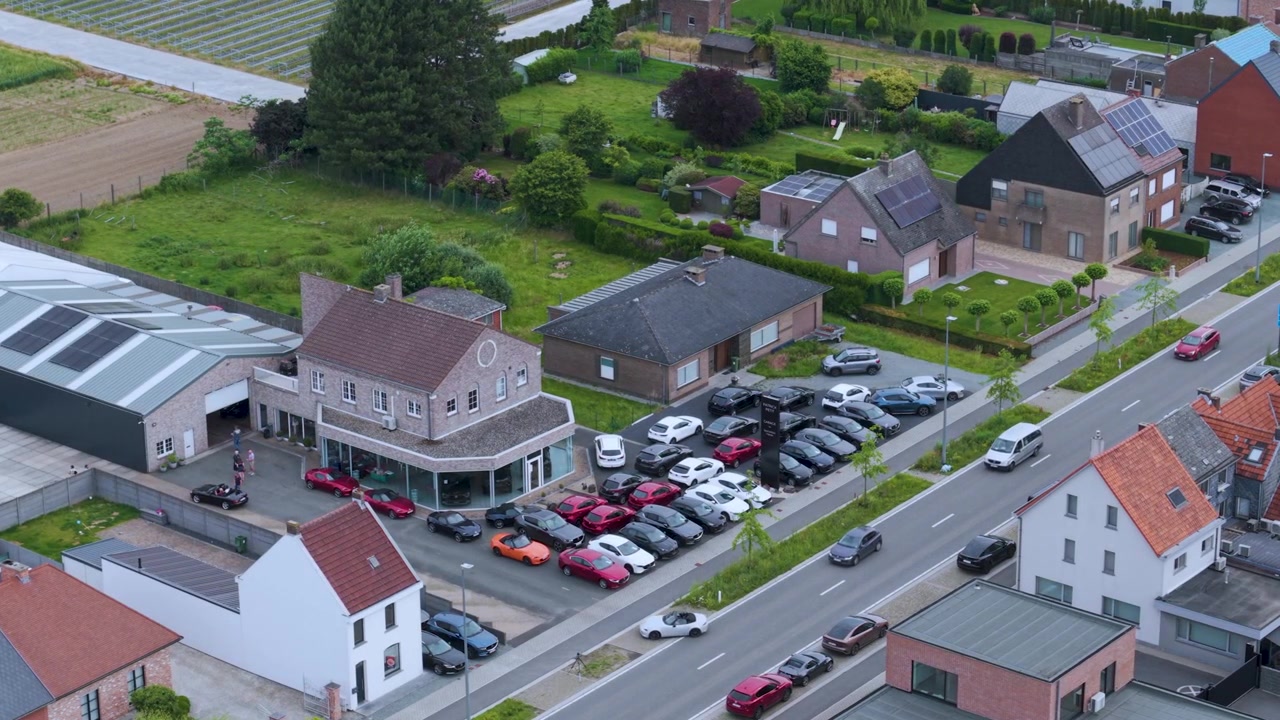 Vista aerea di una concessionaria di auto sportive con file di veicoli parcheggiati, moderni edifici commerciali, accesso stradale e campagna circostante, ripresa in pieno giorno