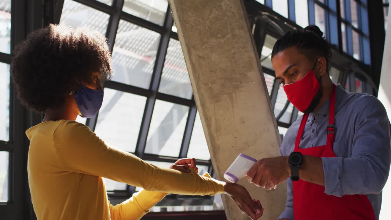 Mixed race male cafe worker wearing face mask taking temperature of female coworker
