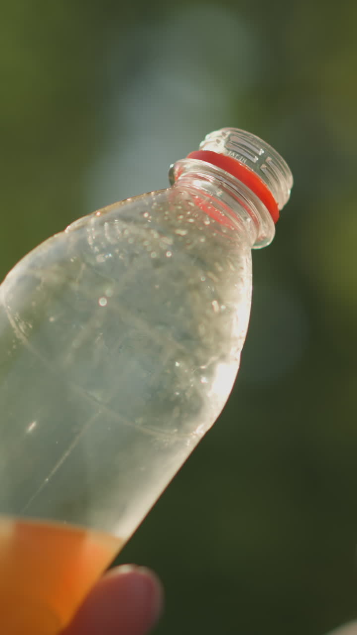 은 플라스틱 병을 들고 있는 손의 클로즈업, 콘덴스와 약간의 액체, 병이 빛을 향해 기울어, 배경에 부드러운 빛을 만들어, 흐릿한 초록색과 함께