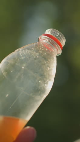 primer plano de la mano sosteniendo una botella de plástico transparente con condensación y algo de líquido, la botella inclinada hacia la luz solar, creando un brillo suave en el fondo, con vegetación borrosa