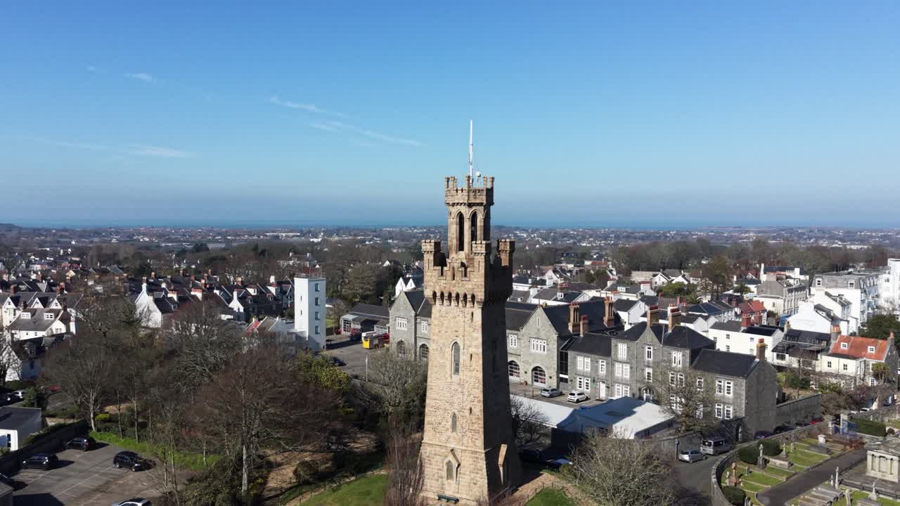 Filmato ravvicinato con drone in circolo di St Peter Port Guernsey, che mostra la torre dell'Arsenale, la stazione dei pompieri e il cimitero, con vista su tutta l'isola fino alla baia di Belle Greve, sotto il sole del tardo pomeriggio e un orizzonte nebbioso.