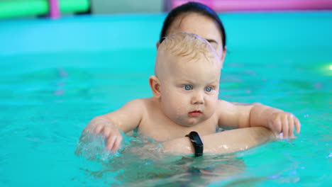 dark-haired woman talks to a baby she's holding in the swimming pool. adorable kid splashes water with two hands. close up.