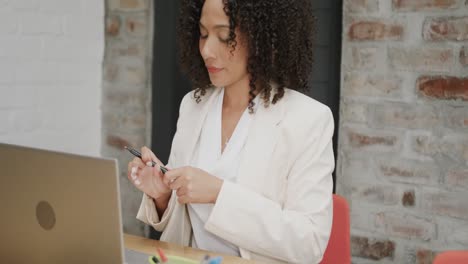 mujer de negocios biracial pensativa sentada en el escritorio con un bolígrafo y usando una computadora portátil, en cámara lenta