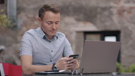 medium shot of young man wearing streetwear sitting in restaurant or cafe in front of laptop and having mobile phone. breakfast with smartphone