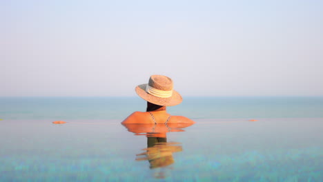 Back-of-Young-female-in-swimsuit-and-summer-sunhat-enjoying-stunning-sunset-view-on-sea-skyline-from-infinity-swimming-pool,-luxury-holiday-concept,-wide-angle