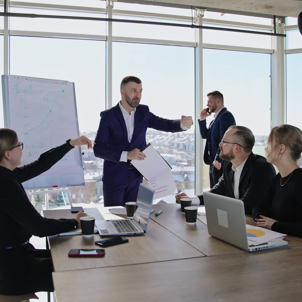 Colleagues sitting at the table in front of laptops and listening to the speaker. Male partner standing at the window and speaking on the phone at the backdrop