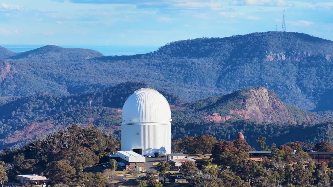 Aerial footage shows a white observatory dome amid forested hills and mountains, with smooth lateral camera movement and warm, natural sunset lighting