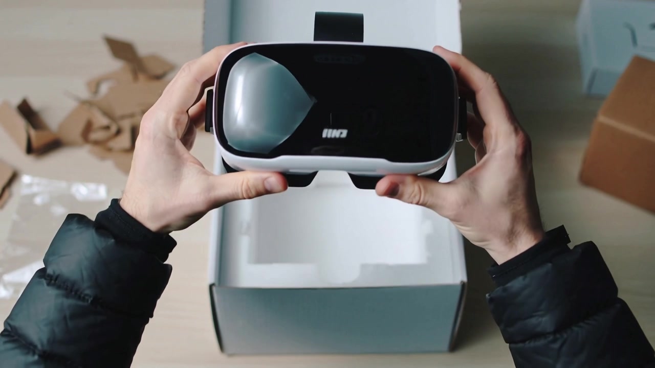 Opening white box, hands in dark jacket lifting VR headset at desk, inspecting alignment for setup