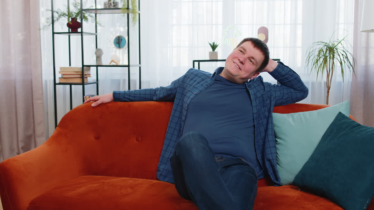 행복한 백인 남자가 집 방에 들어오고 오렌지색 소파에 앉아 휴식을 취하고 꿈을 꾸고 휴식을 취합니다.