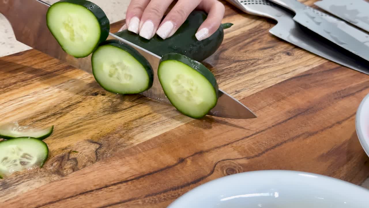 A person uses a chef’s knife to slice a cucumber on a wooden cutting board under bright, even kitchen lighting, with close-up camera angles