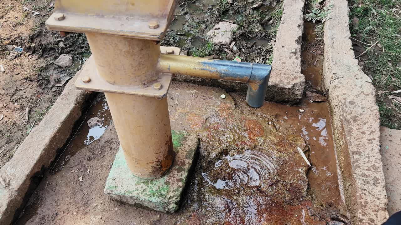 Cirkelend shot van een roestige handpomp met water dat op een stenen voet druppelt, natte betonnen goot, minerale vlekken, landelijke grondtexturen, langzame beweging die het verval en de levendigheid vastlegt.