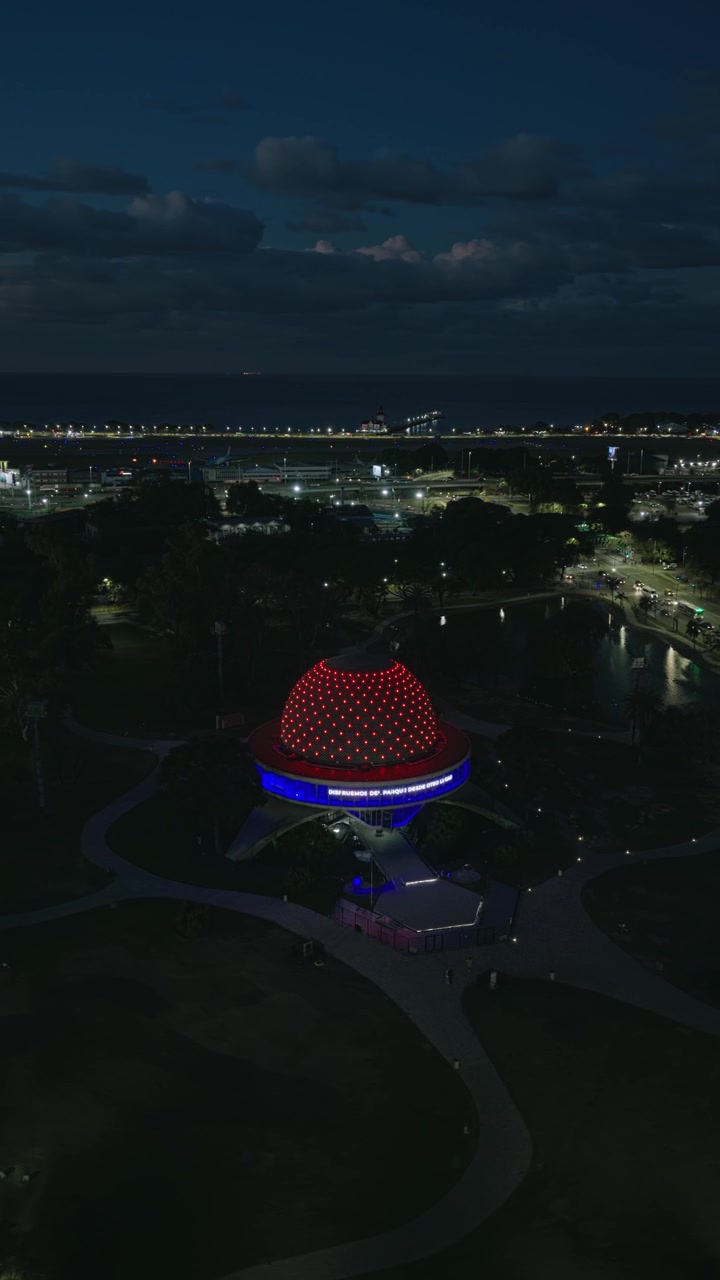 Vertical aerial semi orbit around the illuminated Galileo Galilei Planetarium at night as an airplane passes in the distance.