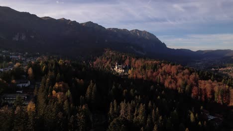 Castillo-De-Peles-En-Medio-De-Un-Bosque-De-Colores-Otoñales-Con-Las-Montañas-De-Bucegi-Como-Telón-De-Fondo-Al-Atardecer,-Vista-Aérea