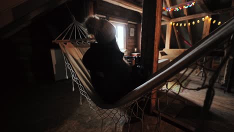 woman in warm coat and hat rocks gently in hammock with drink, cozy attic cabin