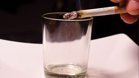 empty glass used for a smoker that is flicking leftover ash into it instead of using ashtray cigarette with golden filter caucasian hand with nails cut very short onto the skin close up slow motion