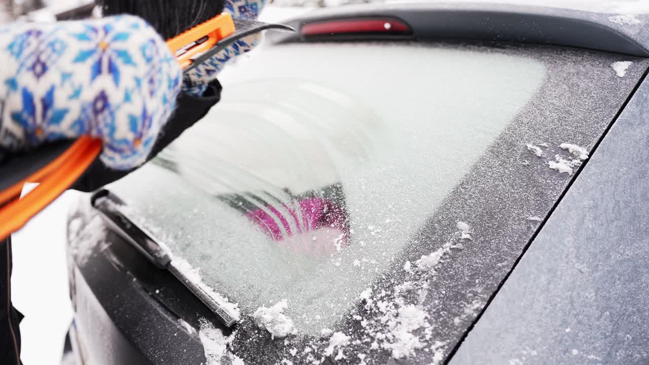 Gloved hands use a brush ice scraper to clear frost from a vehicle’s rear windshield, revealing glass and improving visibility before a winter drive