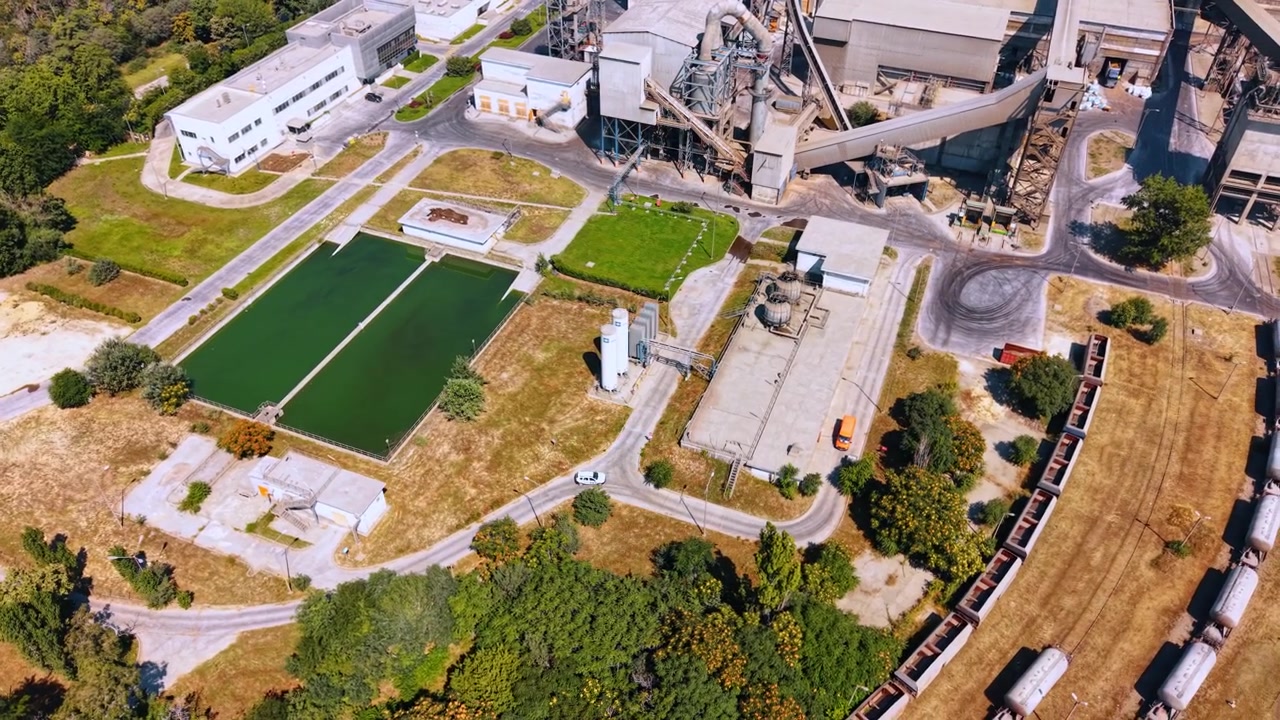 Balsas de decantación industriales junto a una planta de procesamiento de cemento. Vista de dron cenital muestra dos estanques verdes, estructuras de fábrica, caminos de servicio, tanques y vagones de carga.