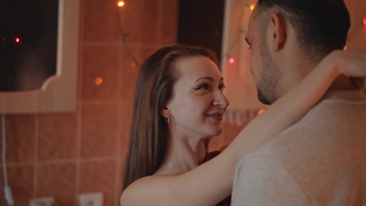 Young couple sharing tender embrace inside warmly lit tiled bathroom, smiling woman leaning into man's shoulder while hand rests gently, creating relaxed intimate domestic mood with soft glow