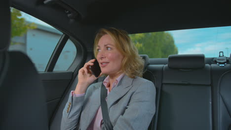 Mature-Businesswoman-Sitting-In-Back-Seat-Of-Taxi-Or-Car-Talking-On-Mobile-Phone