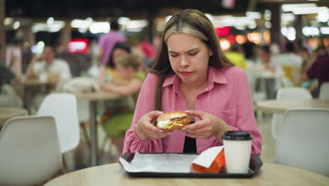 dama de vestido rosa huele hamburguesa con una expresión de descontento, luego lo deja caer, ella alcanza una servilleta para limpiarse la mano, mostrando asco, con vista borrosa de la gente comiendo en el fondo