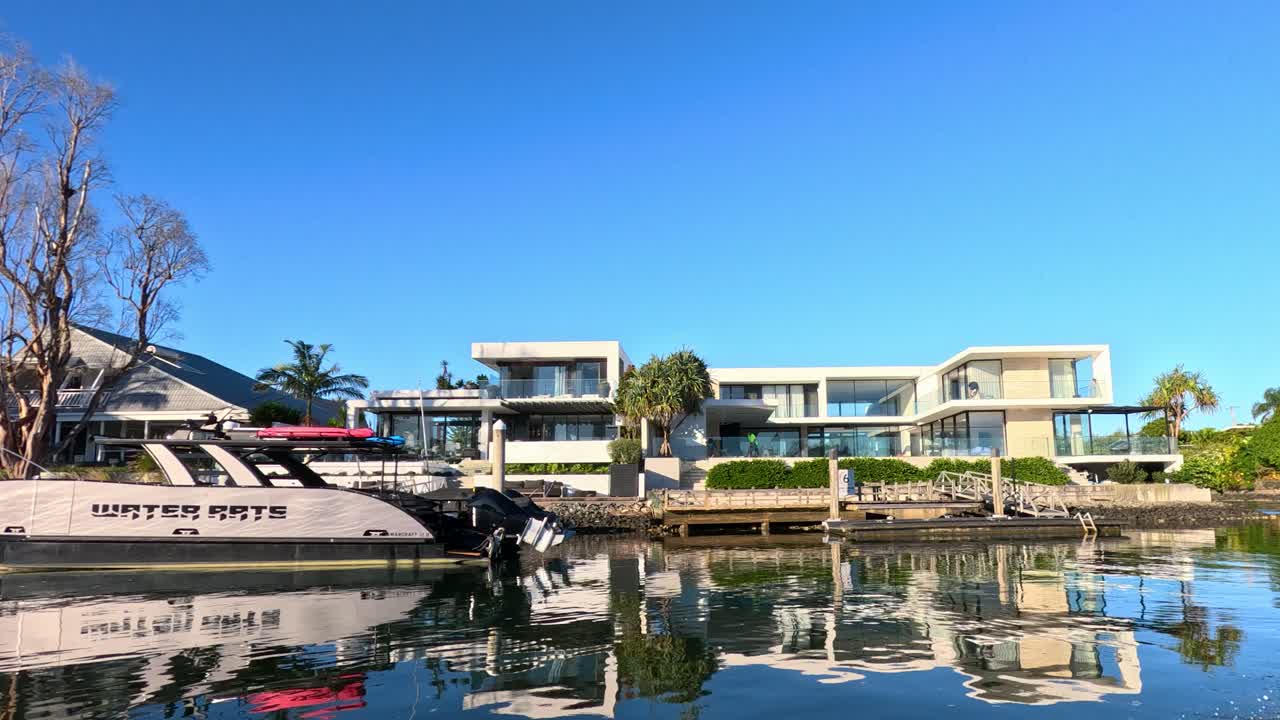A modern luxury house with palm trees is viewed across a calm river as a motorboat smoothly enters the frame, under bright daylight with clear blue skies