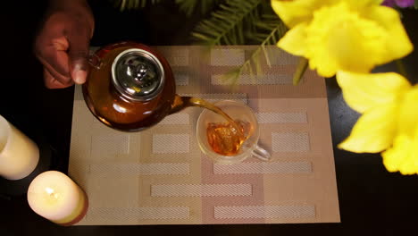 Top-down-shot-of-an-African-American-man-pouring-steaming-hot-tea-into-a-heart-shaped-transparent-glass-tea-cup-sitting-on-a-cozy-dining-table