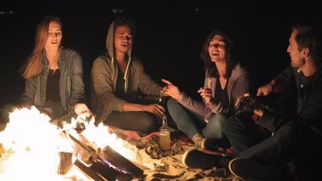 the bonfire late at night: young cheerful people sitting by the fire in the evening, playing guitar and drinking beer. cheerful friends singing songs, talking and having fun together