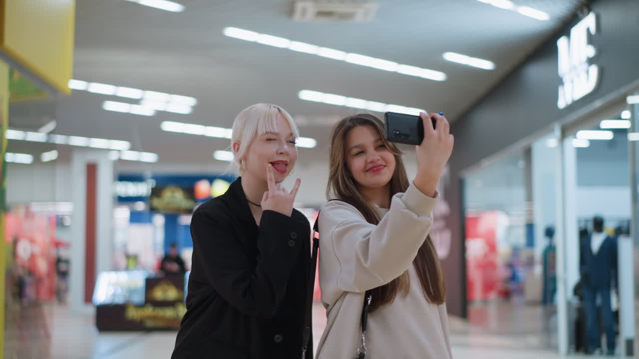 Two teens pose while taking selfie with smartphone inside shopping mall, making playful expressions and standing close together under bright ceiling lights with retail shops visible in background