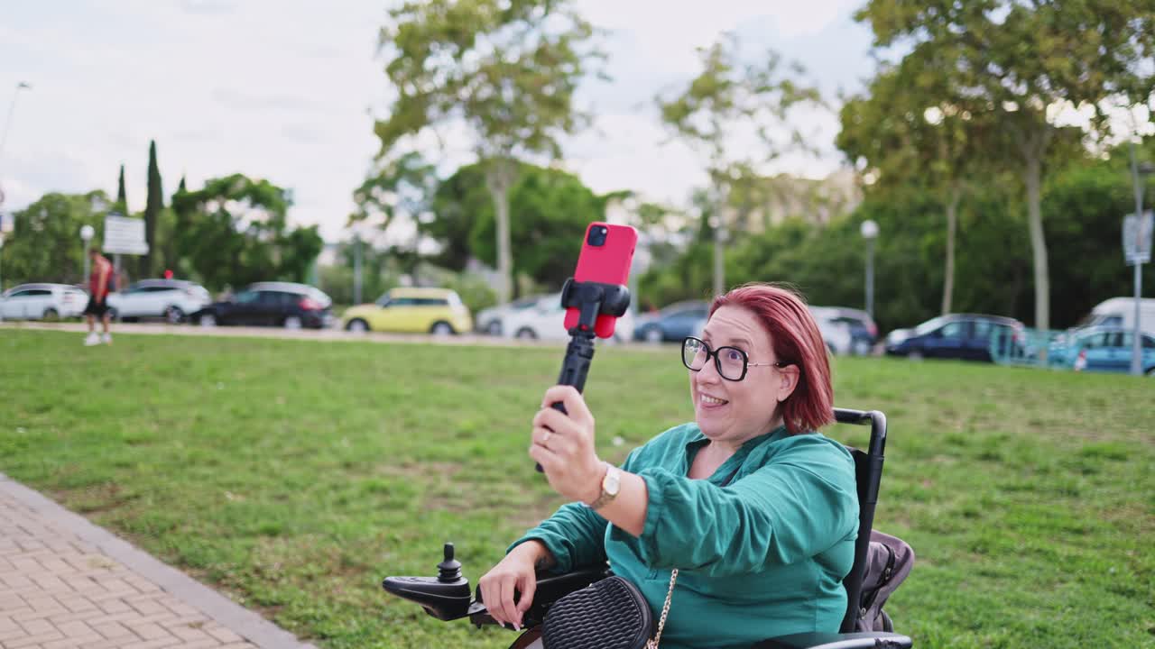Glückliche Frau im Rollstuhl benutzt Smartphone mit Selfie-Stick im Park