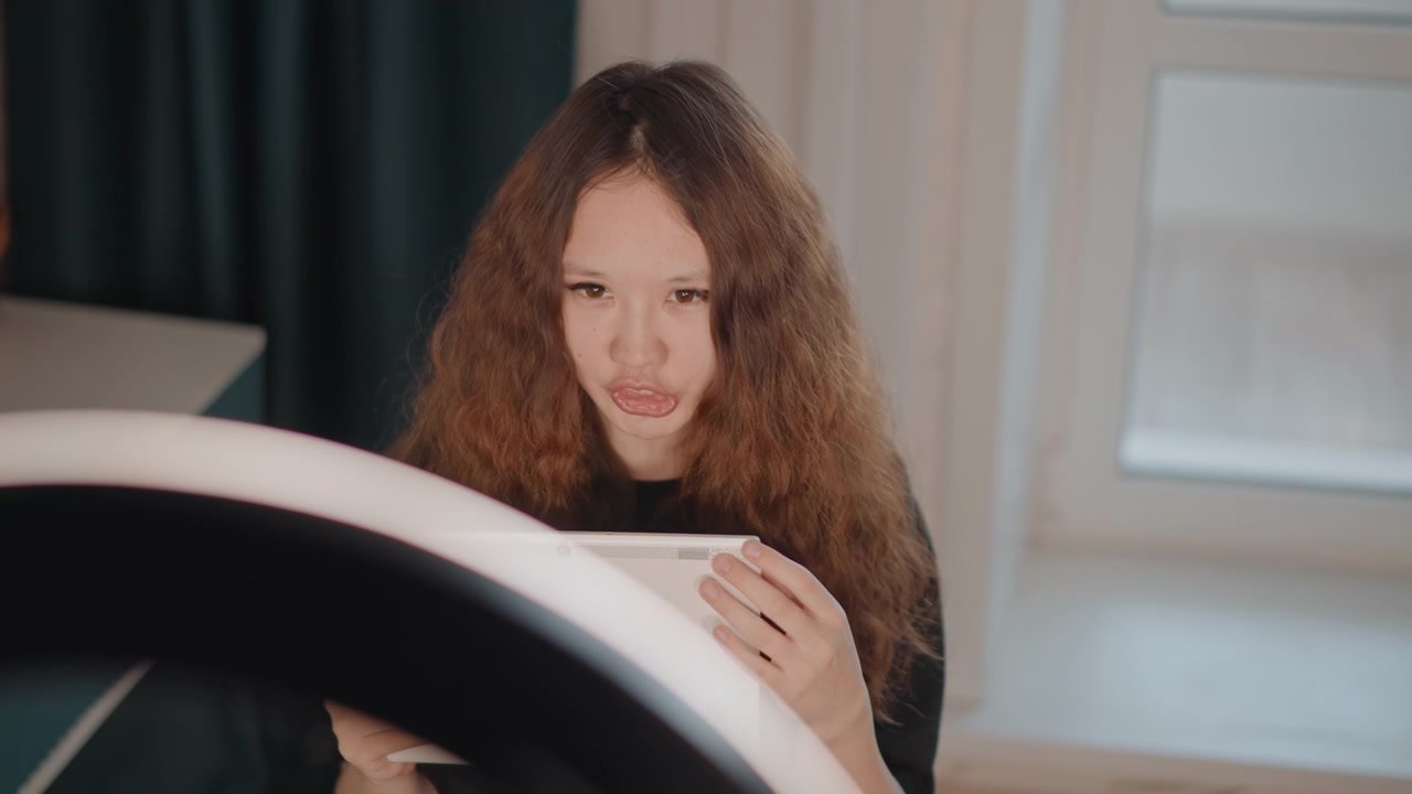 Young woman with long curly hair sits indoors holding tablet, reading screen, then making conversational hand gestures while leaning forward slightly; soft ring light and window light shape calm
