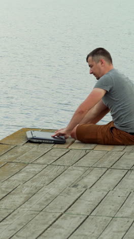 a man is approaching to the end of a masonry and opening a laptop on the background of lake in the summer. Vertical video
