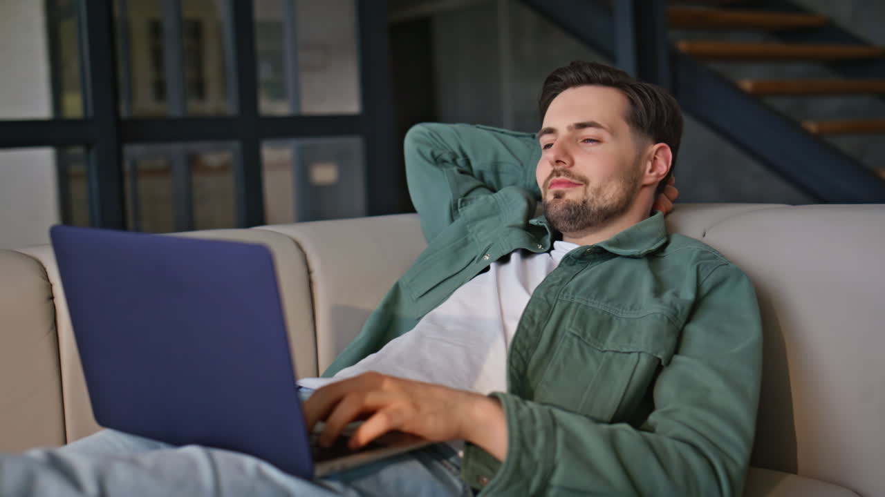 Relaxed guy lounging couch looking laptop screen at house environment closeup