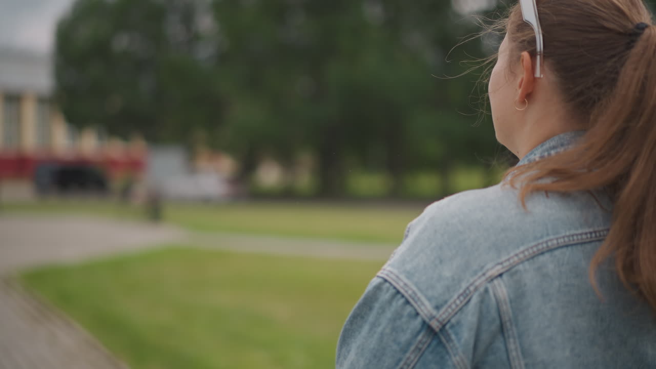 Jovem mulher de jaqueta jeans se afastando em direção a um gramado verde e um caminho, sacola no ombro, óculos de sol na cabeça, fundo com bokeh suave, ritmo lento e reflexivo, clima cinematográfico, luz do fim da tarde