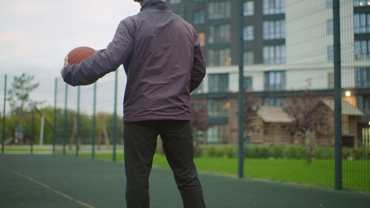 man basketball outdoor court wearing headphones and jacket, solitary athlete rehearsing dribble and warmup moves, urban residential skyline and fenced hoop in soft dusk light, quiet determination