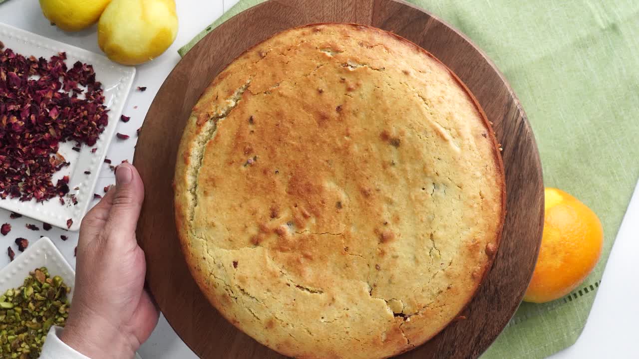 Baking a dessert called Persian Love Cake made with rose petals and pistachios. Cake with white glaze frosting. Putting cake on a wooden plate to cool. Beautiful cake for tea time or celebration