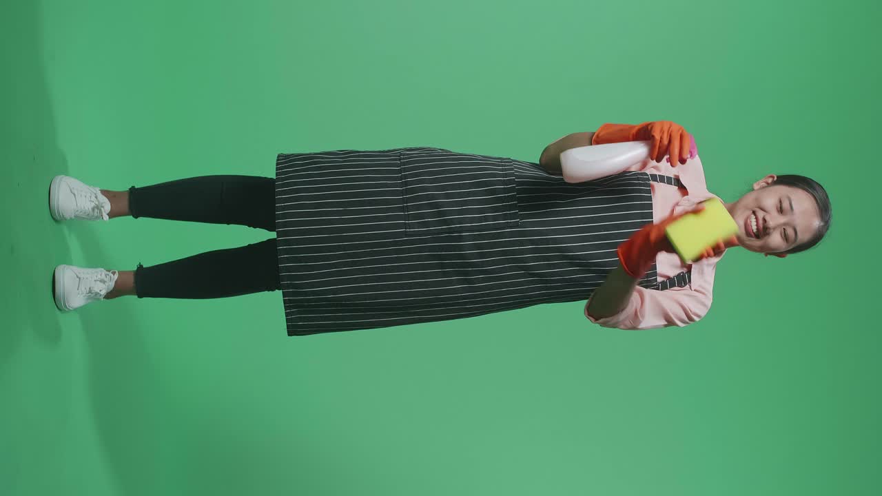 녹색 스크린 스튜디오에서 스프레이와 부 스폰지로 유리잔을 는 아프론을 가진 미소 짓는 아시아 여성 집사.