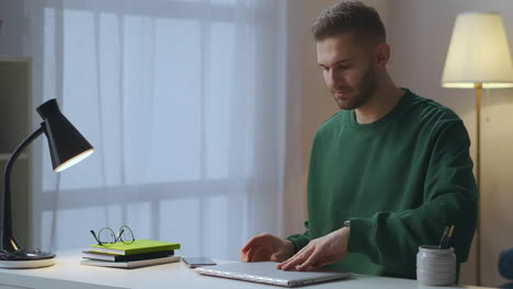 Junger-Gutaussehender-Mann-Im-Grünen-Sweatshirt-Tippt-Auf-Seinem-Laptop,-Internetnutzer-Arbeitet-Allein-In-Seiner-Wohnung,-Stellt-Eine-Verbindung-Zum-Internet-Her-Und-Schaltet-Die-Tischlampe-Ein,-Mittlere-Aufnahme