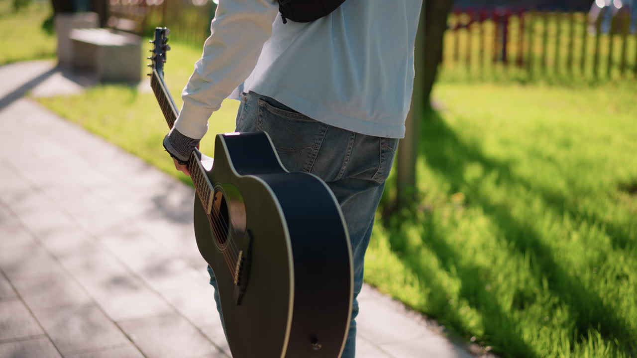 young male guitarist walking through sunlit park, carrying black acoustic case by side wearing jeans and white long sleeve with sling bag, passing bench and stone planter on paved path, warm golden