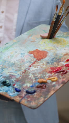 artist draws holding rectangular palette in studio closeup. artworks creator with beard and mustaches draws on on easel canvas. elegant painter at work in shop